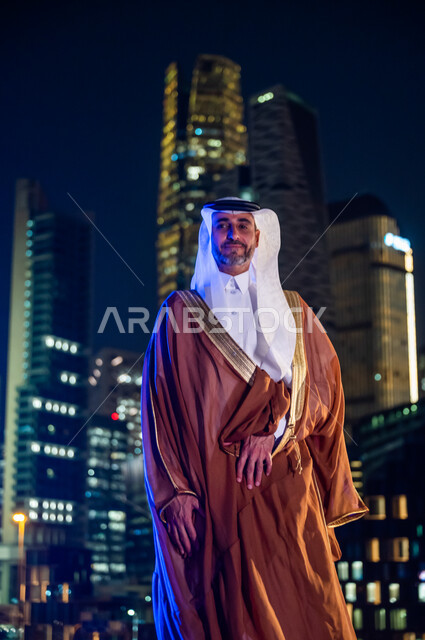 الفخر والاعتزاز بالدولة السعودية, ارتداء الأزياء الشعبية التقليدية، الوقوف بثقة والنظر إلى الكاميرا، الاستمتاع والخروج في الهواء الطلق, رجل عربي خليجي سعودي يرتدي البشت والغترة, خلفية أبراج وناطحات سحاب مدينة الرياض المضاءة في وقت الليل.