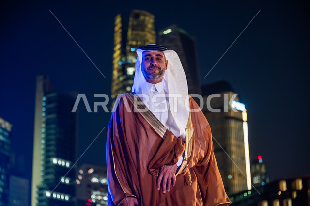 الاعتزا والفخر بالدولة السعودية, ارتداء الأزياء الشعبية التقليدية، الوقوف بثقة والنظر إلى الكاميرا، الاستمتاع والخروج في الهواء الطلق, صورة مقربة لرجل عربي خليجي سعودي يرتدي البشت والغترة, خلفية أبراج وناطحات سحاب مدينة الرياض المضاءة في وقت الليل.