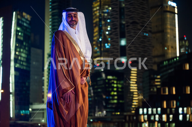Pride and pride in the Saudi state, wearing traditional folk costumes, standing confidently and looking at the camera, having fun and going out in the open air, a Saudi Arabian Gulf man wearing a bisht and ghutra, the background of the towers and skyscrap
