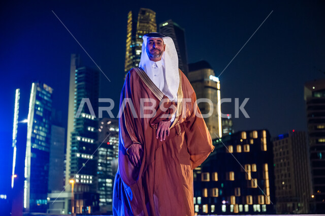 التباهي والفخر بالدولة السعودية, ارتداء الأزياء الشعبية التقليدية، الوقوف بثقة والنظر إلى الكاميرا، الاستمتاع والخروج في الهواء الطلق, رجل عربي خليجي سعودي يرتدي البشت والغترة, خلفية أبراج وناطحات سحاب مدينة الرياض المضاءة في وقت الليل.
