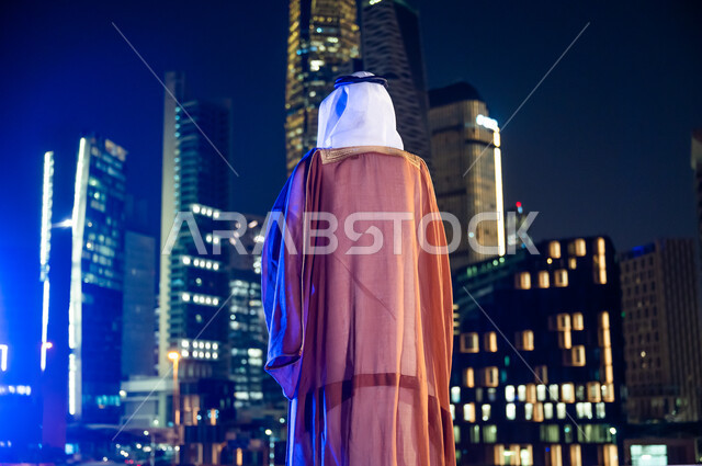 الوقوف لتأمل معالم مدينة الرياض, ارتداء الأزياء الشعبية التقليدية، الاستمتاع والخروج في الهواء الطلق, صورة من الخلف لرجل عربي خليجي سعودي يرتدي البشت والغترة, الأبراج وناطحات السحاب المضاءة في وقت الليل.