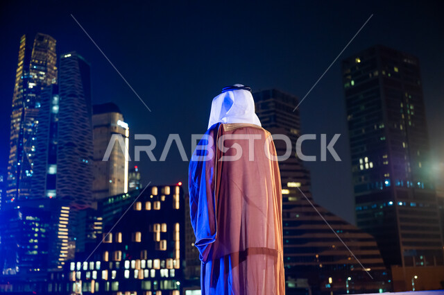 الوقوف لتأمل معالم مدينة الرياض, ارتداء الأزياء الشعبية التقليدية، الاستمتاع والخروج في الهواء الطلق, صورة من الخلف لرجل عربي خليجي سعودي يرتدي البشت والغترة, الأبراج وناطحات السحاب المضاءة في وقت الليل.