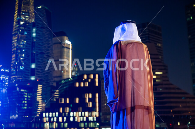 الوقوف لتأمل معالم مدينة الرياض, ارتداء الأزياء الشعبية التقليدية، الاستمتاع والخروج في الهواء الطلق, صورة من الخلف لرجل عربي خليجي سعودي يرتدي البشت والغترة, الأبراج وناطحات السحاب المضاءة في وقت الليل.