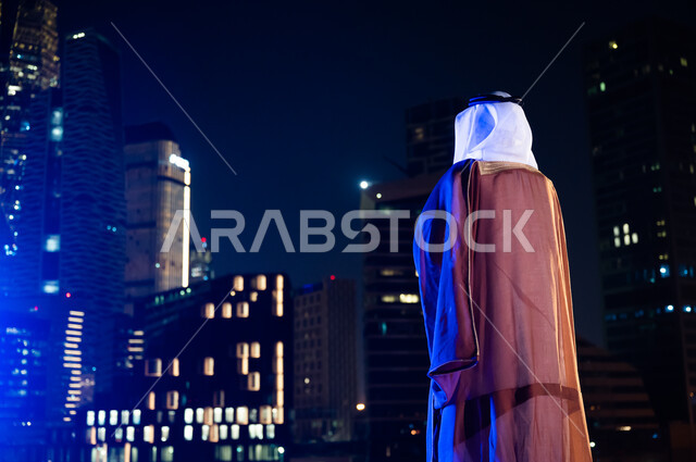 الوقوف لتأمل معالم مدينة الرياض, ارتداء الأزياء الشعبية التقليدية، الاستمتاع والخروج في الهواء الطلق, صورة من الخلف لرجل عربي خليجي سعودي يرتدي البشت والغترة, الأبراج وناطحات السحاب المضاءة في وقت الليل.