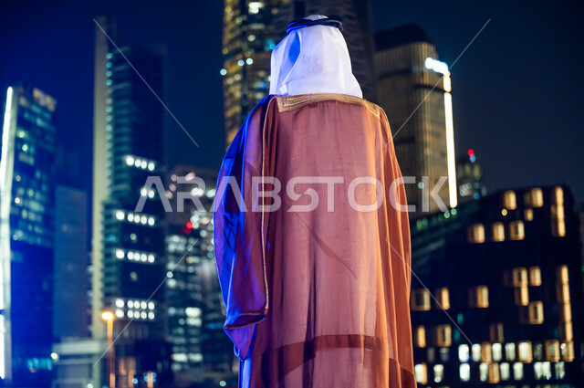 الأبراج وناطحات السحاب المضاءة في وقت الليل، الوقوف لتأمل معالم مدينة الرياض, ارتداء الأزياء الشعبية التقليدية، الاستمتاع والخروج في الهواء الطلق, صورة من الخلف لرجل عربي خليجي سعودي يرتدي البشت والغترة.