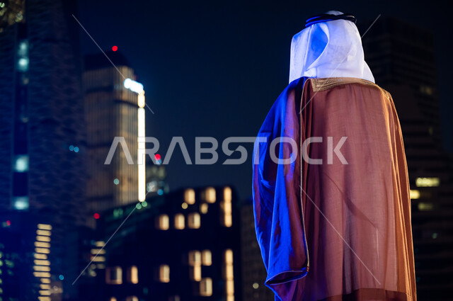 الأبراج وناطحات السحاب المضاءة في وقت الليل، الوقوف لتأمل معالم مدينة الرياض, ارتداء الأزياء الشعبية التقليدية، الاستمتاع والخروج في الهواء الطلق, صورة من الخلف لرجل عربي خليجي سعودي يرتدي البشت والغترة.