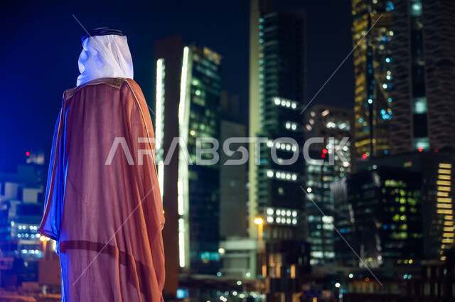 Towers and skyscrapers illuminated at night time, standing to contemplate the landmarks of the city of Riyadh, wearing traditional folk costumes, enjoying and going out in the open air, a picture from behind of a Saudi Arabian Gulf man wearing a bisht and