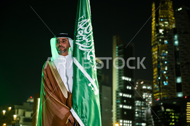 مفهوم حب الوطن والانتماء له، الحفاظ على الهوية الوطنية والافتخار بتراثها, رجل عربي سعودي خليجي يرتدي البشت والغترة يحتفل باليوم الوطني 23 سبتمبر, يوم العلم السعودي 11 مارس, العيد الوطني السعودي, خلفية الابراج وناطحات السحاب في الرياض ليلا.