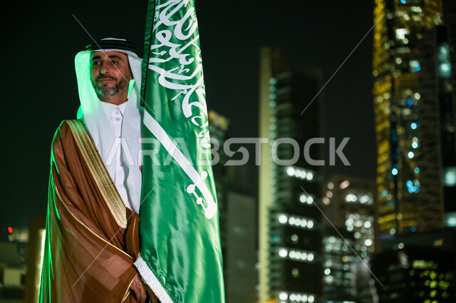 مفهوم حب الوطن والانتماء له، الحفاظ على الهوية الوطنية والافتخار بتراثها, رجل عربي سعودي خليجي يرتدي البشت والغترة يحتفل باليوم الوطني 23 سبتمبر, يوم العلم السعودي 11 مارس, العيد الوطني السعودي, خلفية الابراج وناطحات السحاب في الرياض ليلا.