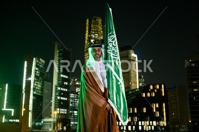 الوقوف بثقة وفخر والنظر إلى الكاميرا، أعياد ومناسبات وطنية، رجل عربي سعودي خليجي يرتدي البشت والغترة يحتفل باليوم الوطني 23 سبتمبر, يوم العلم السعودي 11 مارس, العيد الوطني السعودي, خلفية مركز الملك عبد الله المالي(كافد) في الرياض ليلا.