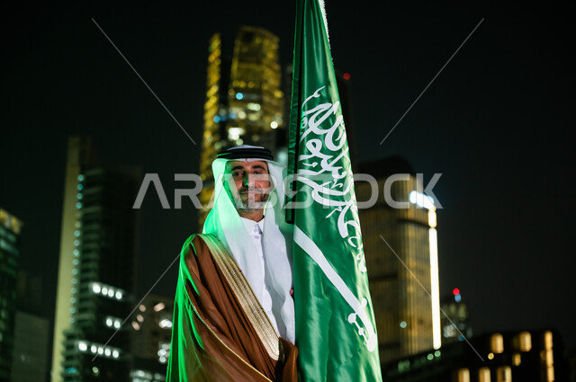 الوقوف بثقة وفخر والنظر إلى الكاميرا، أعياد ومناسبات وطنية، رجل عربي سعودي خليجي يرتدي البشت والغترة يحتفل باليوم الوطني 23 سبتمبر, يوم العلم السعودي 11 مارس, العيد الوطني السعودي, خلفية مركز الملك عبد الله المالي(كافد) في الرياض ليلا.
