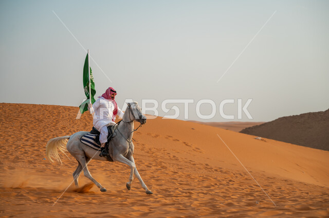 العيد الوطني 23 سبتمبر، يوم العلم السعودي 11 مارس، علاقة أبناء الوطن بالخيل السعودي الأصيل، رجل عربي خليجي سعودي يرتدي الشماغ والزي التقليدي يمتطي حصانه يمسك بيده العلم في الصحراء وقت غروب الشمس، الإحتفال بذكرى اليوم الوطني السعودي