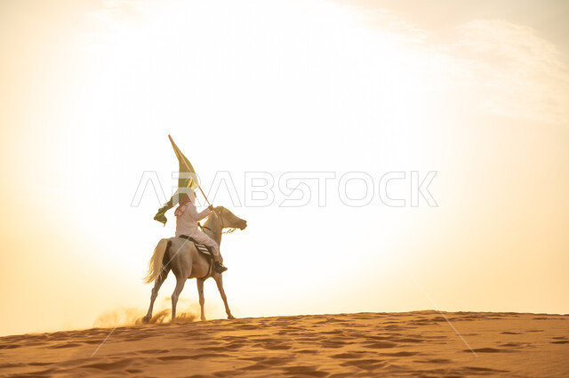 العيد الوطني 23 سبتمبر، يوم العلم السعودي 11 مارس، علاقة أبناء الوطن بالخيل السعودي الأصيل، رجل عربي خليجي سعودي يرتدي الشماغ والزي التقليدي يمتطي حصانه يمسك بيده العلم في الصحراء وقت غروب الشمس، الإحتفال بذكرى اليوم الوطني السعودي