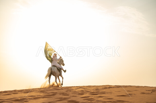 العيد الوطني 23 سبتمبر، يوم العلم السعودي 11 مارس، علاقة أبناء الوطن بالخيل السعودي الأصيل، رجل عربي خليجي سعودي يرتدي الشماغ والزي التقليدي يمتطي حصانه يمسك بيده العلم في الصحراء وقت غروب الشمس، الإحتفال بذكرى اليوم الوطني السعودي