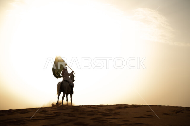العيد الوطني 23 سبتمبر، يوم العلم السعودي 11 مارس، علاقة أبناء الوطن بالخيل السعودي الأصيل، رجل عربي خليجي سعودي يرتدي الشماغ والزي التقليدي يمتطي حصانه يمسك بيده العلم في الصحراء وقت غروب الشمس، الإحتفال بذكرى اليوم الوطني السعودي