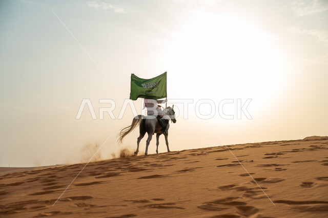 الإحتفال بذكرى اليوم الوطني 23 سبتمبر ، العيد الوطني السعودي، يوم العلم 11 مارس، علاقة أبناء الوطن بالخيل السعودي الأصيل، رجل عربي خليجي سعودي يرتدي الشماغ والزي التقليدي يمتطي حصانه يمسك بيده العلم في الصحراء وقت غروب الشمس
