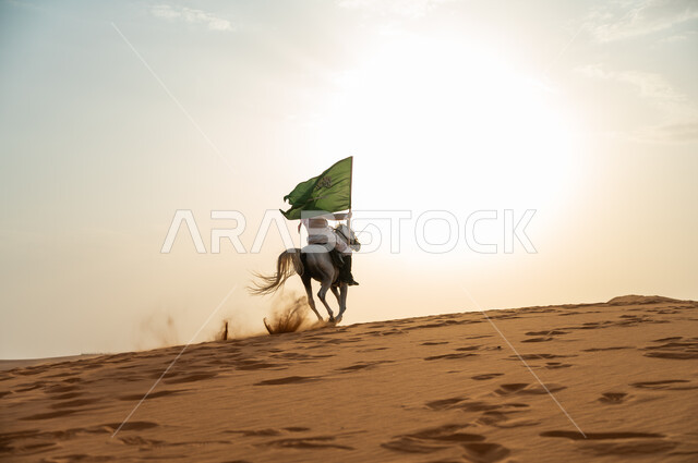 الإحتفال بذكرى اليوم الوطني 23 سبتمبر ، العيد الوطني السعودي، يوم العلم 11 مارس، علاقة أبناء الوطن بالخيل السعودي الأصيل، رجل عربي خليجي سعودي يرتدي الشماغ والزي التقليدي يمتطي حصانه يمسك بيده العلم في الصحراء وقت غروب الشمس