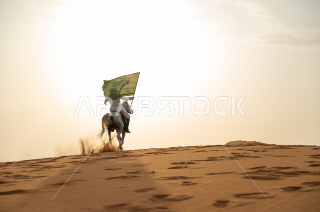 الإحتفال بذكرى اليوم الوطني 23 سبتمبر ، العيد الوطني السعودي، يوم العلم 11 مارس، علاقة أبناء الوطن بالخيل السعودي الأصيل، رجل عربي خليجي سعودي يرتدي الشماغ والزي التقليدي يمتطي حصانه يمسك بيده العلم في الصحراء وقت غروب الشمس