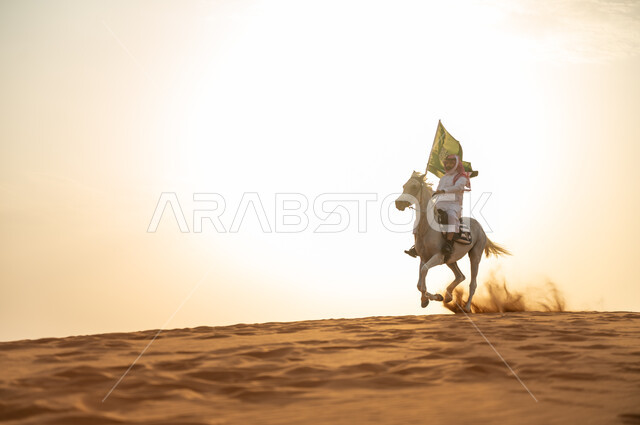 الإحتفال بذكرى اليوم الوطني 23 سبتمبر ، العيد الوطني السعودي، يوم العلم 11 مارس، علاقة أبناء الوطن بالخيل السعودي الأصيل، رجل عربي خليجي سعودي يرتدي الشماغ والزي التقليدي يمتطي حصانه يمسك بيده العلم في الصحراء وقت غروب الشمس