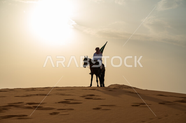 احياء ذكرى اليوم الوطني السعودي 23 سبتمبر، الاحتفال بيوم العلم السعودي 11 مارس، علاقة أبناء الوطن بالخيل السعودي الأصيل، رجل عربي خليجي سعودي يرتدي الشماغ والزي التقليدي يمتطي حصانه يمسك بيده العلم في الصحراء وقت غروب الشمس