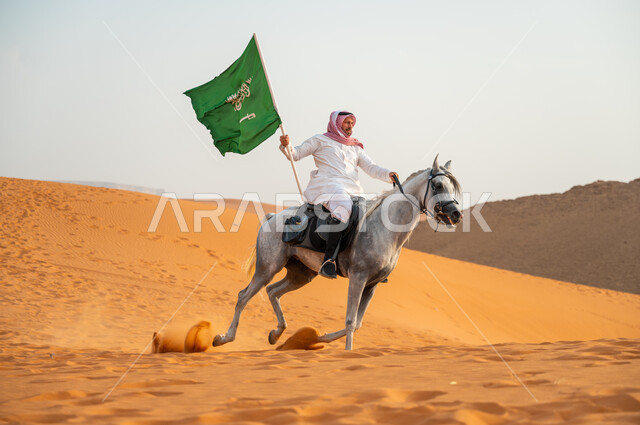 احياء ذكرى اليوم الوطني السعودي 23 سبتمبر، الاحتفال بيوم العلم السعودي 11 مارس، علاقة أبناء الوطن بالخيل السعودي الأصيل، رجل عربي خليجي سعودي يرتدي الشماغ والزي التقليدي يمتطي حصانه يمسك بيده العلم في الصحراء وقت غروب الشمس