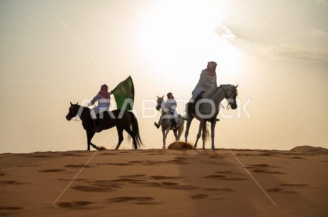 أنشطة وفعاليات ركوب الجياد في الصحراء، ذكرى اليوم الوطني السعودي 23 سبتمبر، رفع الراية للاحتفال بيوم العلم السعودي 11 مارس، مجموعة رجال عرب خليجيون سعوديون يتجولون على ظهر الاحصنة وقت غروب الشمس، حب وعشق الخيل السعودي الأصيل