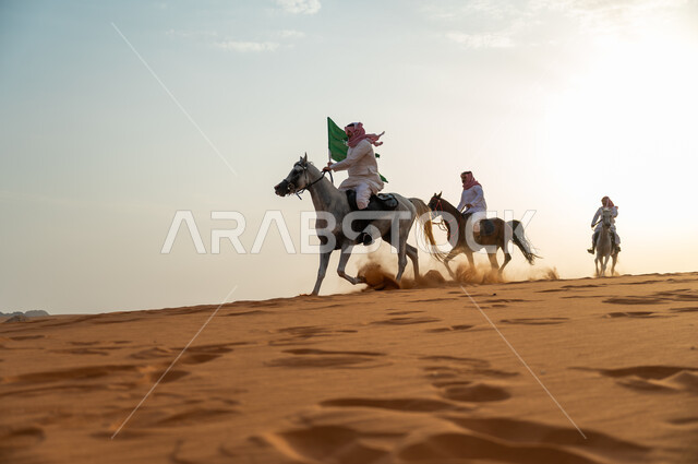 أنشطة وفعاليات ركوب الجياد في الصحراء، ذكرى اليوم الوطني السعودي 23 سبتمبر، رفع الراية للاحتفال بيوم العلم السعودي 11 مارس، مجموعة رجال عرب خليجيون سعوديون يتجولون على ظهر الاحصنة وقت غروب الشمس، حب وعشق الخيل السعودي الأصيل