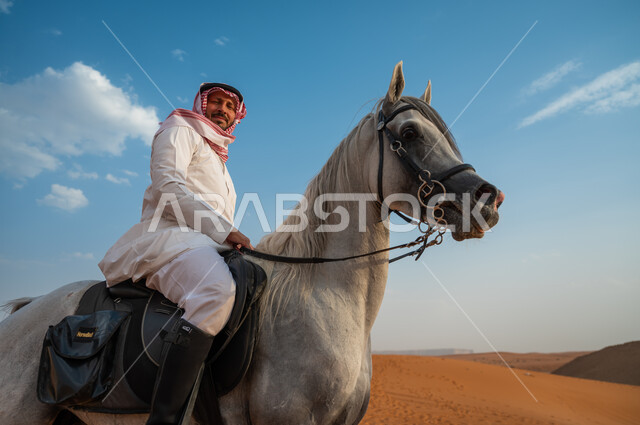 شغف الفروسية وممارسة هواية ركوب الخيل، صورة مقربة لخيال عربي خليجي سعودي مبتسم يرتدي الشماغ والزي التقليدي يركب على حصانه وسط الطبيعه الصحراوية وضح النهار، النظر الى الكاميرا، اتباع تقاليد الأجداد برعاية وتربية الجياد، فعاليات امتطاء الأحصنة في الصحراء
