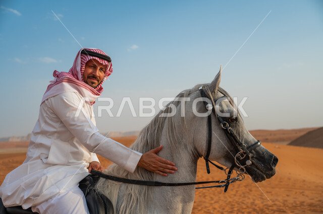 شغف الفروسية وممارسة هواية ركوب الخيل، صورة مقربة لخيال عربي خليجي سعودي مبتسم يرتدي الشماغ والزي التقليدي يركب على حصانه وسط الطبيعه الصحراوية وضح النهار، النظر الى الكاميرا، اتباع تقاليد الأجداد برعاية وتربية الجياد، فعاليات امتطاء الأحصنة في الصحراء