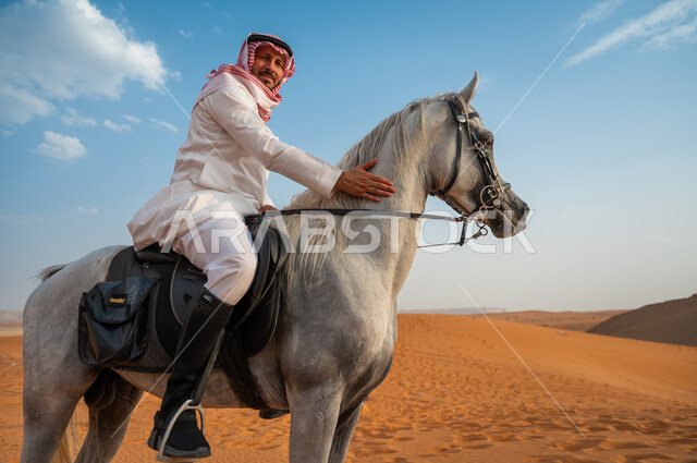 شغف الفروسية وممارسة هواية ركوب الخيل، صورة مقربة لخيال عربي خليجي سعودي مبتسم يرتدي الشماغ والزي التقليدي يركب على حصانه وسط الطبيعه الصحراوية وضح النهار، النظر الى الكاميرا، اتباع تقاليد الأجداد برعاية وتربية الجياد، فعاليات امتطاء الأحصنة في الصحراء