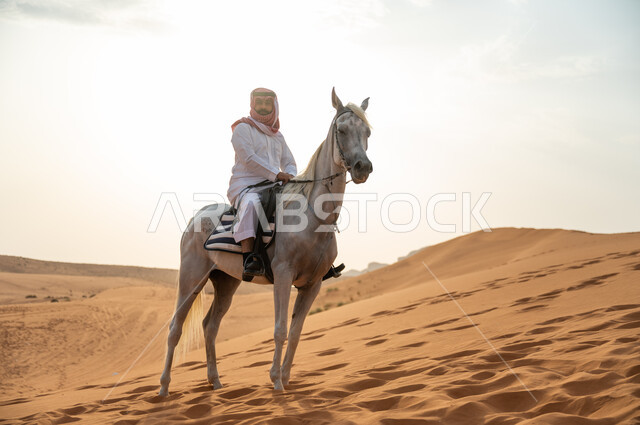 ممارسة هواية ركوب الخيل، رجل عربي خليجي سعودي يرتدي الشماغ والزي التقليدي يركب على حصانه الابيض وسط الطبيعه الصحراوية وضح النهار، فعاليات امتطاء الأحصنة في الصحراء، شغف الفروسية وحب الجواد السعودي الاصيل، اتباع تقاليد الأجداد برعاية وتربية الجياد