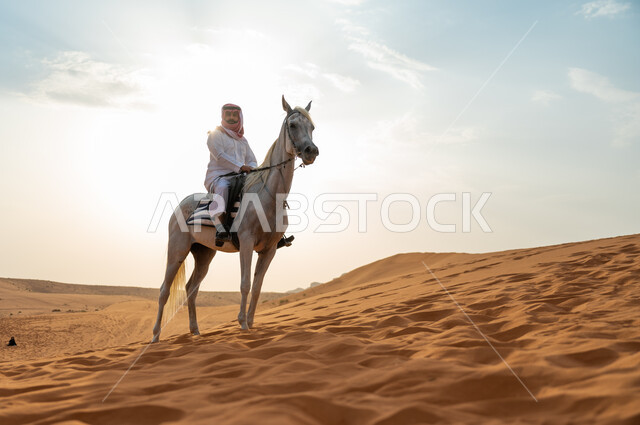 ممارسة هواية ركوب الخيل، رجل عربي خليجي سعودي يرتدي الشماغ والزي التقليدي يركب على حصانه الابيض وسط الطبيعه الصحراوية وضح النهار، فعاليات امتطاء الأحصنة في الصحراء، شغف الفروسية وحب الجواد السعودي الاصيل، اتباع تقاليد الأجداد برعاية وتربية الجياد