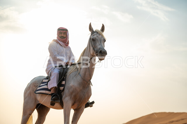 ممارسة هواية ركوب الخيل، رجل عربي خليجي سعودي يرتدي الشماغ والزي التقليدي يركب على حصانه الابيض وسط الطبيعه الصحراوية وضح النهار، فعاليات امتطاء الأحصنة في الصحراء، شغف الفروسية وحب الجواد السعودي الاصيل، اتباع تقاليد الأجداد برعاية وتربية الجياد