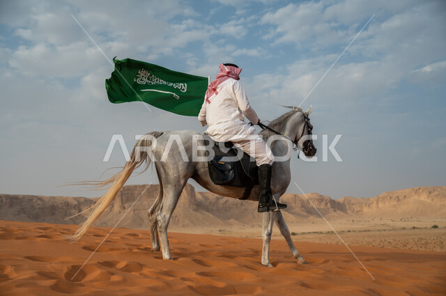 الفخر والتباهي في الإنتماء للسعودية، التجول في الصحراء على ظهر الحصان، فارس عربي خليجي سعودي يرتدي الشماغ والزي التقليدي يمتطي خيل عربي أصيل يرفع علم السعودية، الإحتفال باليوم الوطني السعودي، ذكرى يوم العلم 11 مارس، العيد الوطني لتوحيد البلاد 8 ربيع الأول