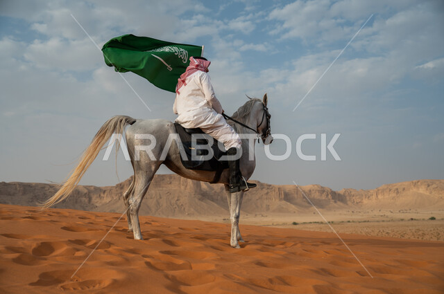 الفخر والتباهي في الإنتماء للسعودية، التجول في الصحراء على ظهر الحصان، فارس عربي خليجي سعودي يرتدي الشماغ والزي التقليدي يمتطي خيل عربي أصيل يرفع علم السعودية، الإحتفال باليوم الوطني السعودي، ذكرى يوم العلم 11 مارس، العيد الوطني لتوحيد البلاد 8 ربيع الأول