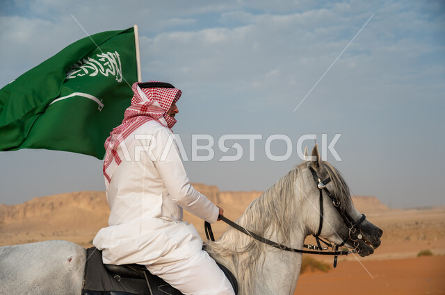 الفخر والتباهي في الإنتماء للسعودية، التجول في الصحراء على ظهر الحصان، فارس عربي خليجي سعودي يرتدي الشماغ والزي التقليدي يمتطي خيل عربي أصيل يرفع علم السعودية، الإحتفال باليوم الوطني السعودي، ذكرى يوم العلم 11 مارس، العيد الوطني لتوحيد البلاد 8 ربيع الأول