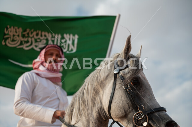 الاعتزاز في الإنتماء للسعودية، التجول على ظهر الحصان وضح النهار، صورة مقربة لرجل عربي خليجي سعودي يرتدي الزي التقليدي يمتطي خيل عربي أصيل يرفع علم السعودية، الإحتفال باليوم الوطني السعودي 23 سبتمبر، ذكرى يوم العلم 11 مارس، العيد الوطني لتوحيد البلاد