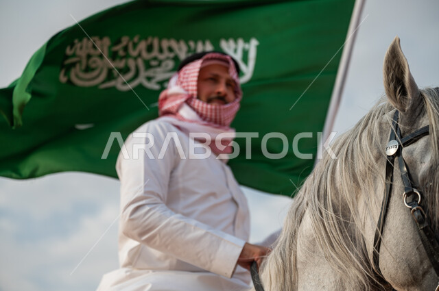 الاعتزاز في الإنتماء للسعودية، التجول على ظهر الحصان وضح النهار، صورة مقربة لرجل عربي خليجي سعودي يرتدي الزي التقليدي يمتطي خيل عربي أصيل يرفع علم السعودية، الإحتفال باليوم الوطني السعودي 23 سبتمبر، ذكرى يوم العلم 11 مارس، العيد الوطني لتوحيد البلاد