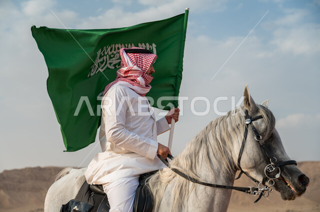 الاعتزاز في الإنتماء للسعودية، التجول على ظهر الحصان وضح النهار، صورة مقربة لرجل عربي خليجي سعودي يرتدي الزي التقليدي يمتطي خيل عربي أصيل يرفع علم السعودية، الإحتفال باليوم الوطني السعودي 23 سبتمبر، ذكرى يوم العلم 11 مارس، العيد الوطني لتوحيد البلاد