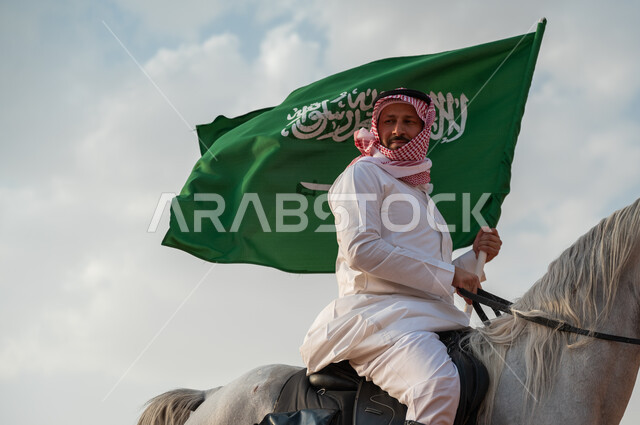 الاعتزاز في الإنتماء للسعودية، التجول على ظهر الحصان وضح النهار، صورة مقربة لرجل عربي خليجي سعودي يرتدي الزي التقليدي يمتطي خيل عربي أصيل يرفع علم السعودية، الإحتفال باليوم الوطني السعودي 23 سبتمبر، ذكرى يوم العلم 11 مارس، العيد الوطني لتوحيد البلاد