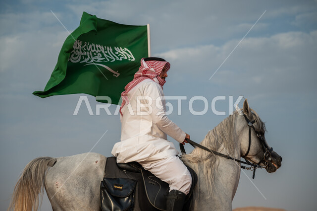 العيد الوطني لتوحيد البلاد، الاعتزاز بالإنتماء للسعودية، التجول على ظهر الحصان وضح النهار، خيال عربي خليجي سعودي يرتدي الشماغ والزي التقليدي يمتطي خيل عربي أصيل يرفع علم السعودية، الإحتفال باليوم الوطني السعودي 23 سبتمبر، ذكرى يوم العلم 11 مارس