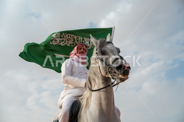 العيد الوطني لتوحيد البلاد، الاعتزاز بالإنتماء للسعودية، التجول على ظهر الحصان وضح النهار، خيال عربي خليجي سعودي يرتدي الشماغ والزي التقليدي يمتطي خيل عربي أصيل يرفع علم السعودية، الإحتفال باليوم الوطني السعودي 23 سبتمبر، ذكرى يوم العلم 11 مارس