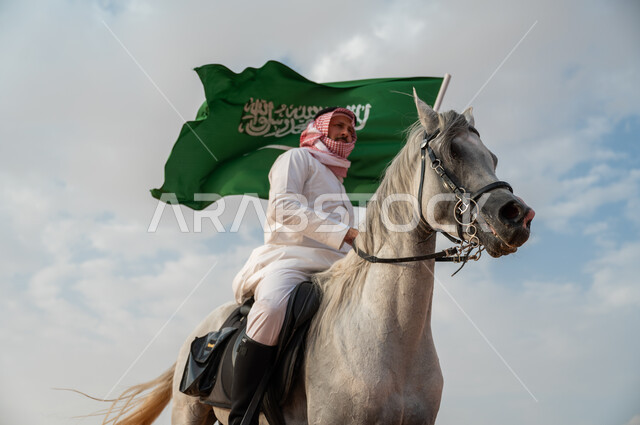 العيد الوطني لتوحيد البلاد، الاعتزاز بالإنتماء للسعودية، التجول على ظهر الحصان وضح النهار، خيال عربي خليجي سعودي يرتدي الشماغ والزي التقليدي يمتطي خيل عربي أصيل يرفع علم السعودية، الإحتفال باليوم الوطني السعودي 23 سبتمبر، ذكرى يوم العلم 11 مارس