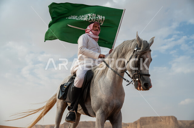 العيد الوطني لتوحيد البلاد، الاعتزاز بالإنتماء للسعودية، التجول على ظهر الحصان وضح النهار، خيال عربي خليجي سعودي يرتدي الشماغ والزي التقليدي يمتطي خيل عربي أصيل يرفع علم السعودية، الإحتفال باليوم الوطني السعودي 23 سبتمبر، ذكرى يوم العلم 11 مارس