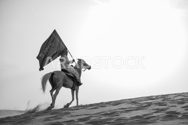 العيد الوطني 23 سبتمبر، يوم العلم السعودي 11 مارس، علاقة أبناء الوطن بالخيل السعودي الأصيل، صورة بالاسود والابيض لرجل عربي خليجي سعودي يرتدي الشماغ والزي التقليدي بمتطي حصانه يمسك بيده العلم في الصحراء، الإحتفال بذكرى اليوم الوطني السعودي
