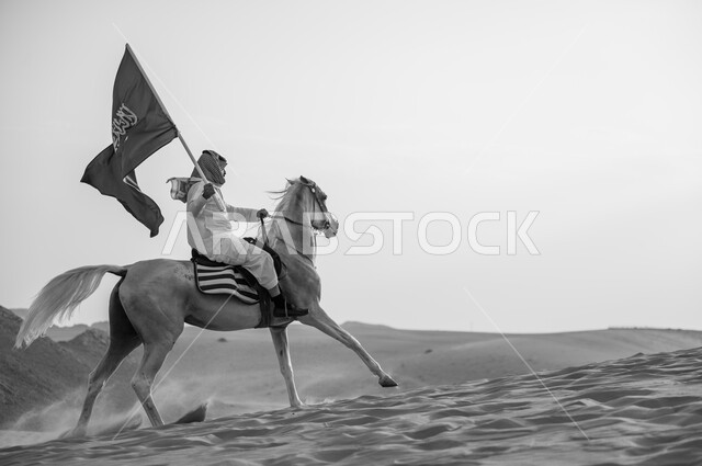 العيد الوطني 23 سبتمبر، يوم العلم السعودي 11 مارس، علاقة أبناء الوطن بالخيل السعودي الأصيل، صورة بالاسود والابيض لرجل عربي خليجي سعودي يرتدي الشماغ والزي التقليدي بمتطي حصانه يمسك بيده العلم في الصحراء، الإحتفال بذكرى اليوم الوطني السعودي