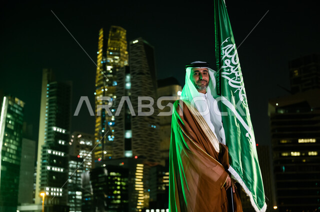 الوقوف باعتزاز وفخر والنظر إلى الكاميرا، أعياد ومناسبات وطنية تقليدية، رجل عربي سعودي خليجي يرتدي البشت والغترة يحتفل باليوم الوطني السعودي , يوم العلم السعودي 11 مارس, العيد الوطني السعودي 8 ربيع الأول , خلفية الأبراج المالية الاقتصادية في الرياض ليلا.