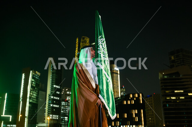 التباهي بالانتماء إلى الدولة السعودية، أعياد ومناسبات وطنية تقليدية، رجل عربي سعودي خليجي يرتدي البشت والغترة يقبل العلم بفخر واعتزاز, يوم العلم السعودي 11 مارس, الاحتفال بذكرى اليوم الوطني السعودي 8 ربيع الأول, خلفية المركز المالي(كافد) في وقت الليل.