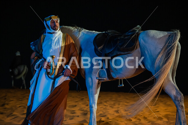 خلفية الطبيعه الرملية الذهبية الناعمة، مشاركة الجياد في مناسية ,يوم التأسيس 1727 م ، حب الفروسيه وركوب الخيل,  رجل سعودي يرتدي البشت والغتره التقليدية يقف بجانب فرسه في الصحراء, ذكرى تأسيس المملكة 22 فبراير