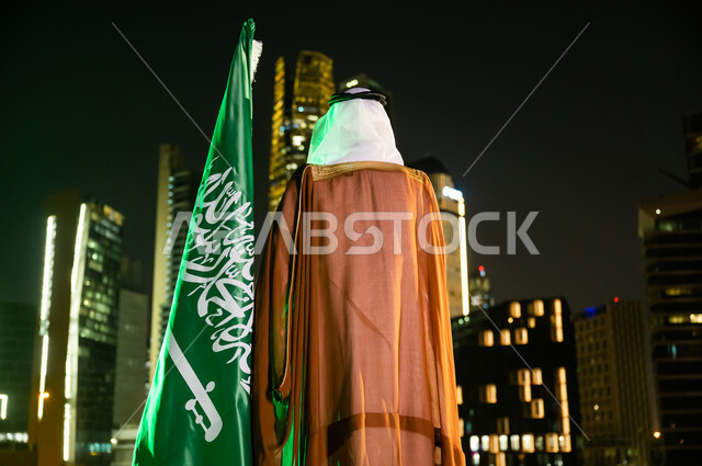 احياء ذكرى اليوم الوطني السعودي 23 سبتمبر،  الاحتفال بيوم العلم السعودي 11 مارس، الوقوف لتأمل الأبراج وناطحات السحاب المضاءة ليلا في مدينة الرياض، صورة من الخلف لرجل عربي خليجي سعودي يرتدي الغترة والبشت يقف بجانب العلم.